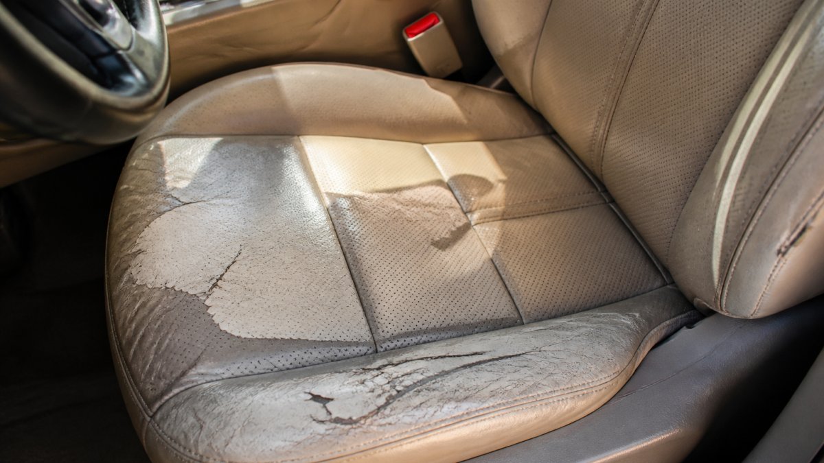 Close-up of a car's leather driver seat showing visible fading, cracking, and peeling from sun exposure, with uneven color patches and surface deterioration clearly visible under bright sunlight